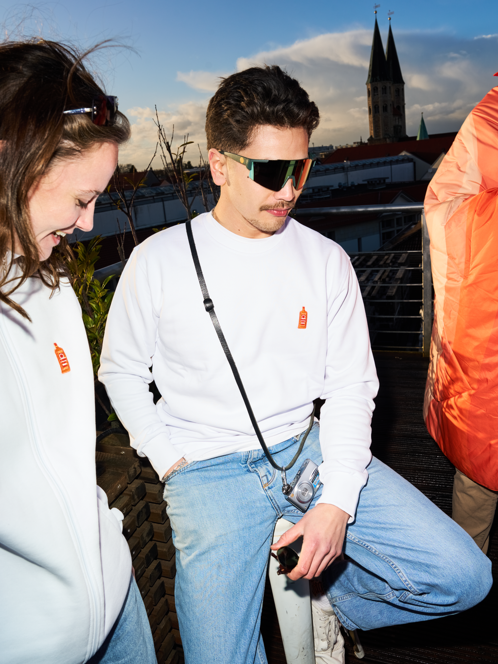 Két ember fehér Jägermeister pulóvert és Jägermeister gyorsszemüveget visel egy tetőteraszon, a háttérben templomtoronnyal és városképpel.