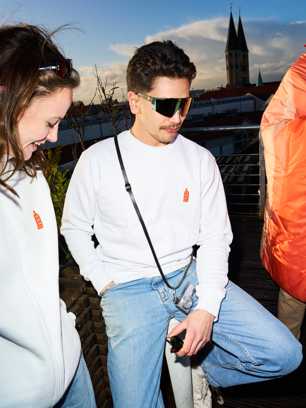 Két ember fehér Jägermeister pulóvert és Jägermeister gyorsszemüveget visel egy tetőteraszon, a háttérben templomtoronnyal és városképpel.