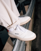 A person wearing off-white Jägermeister sneakers with white socks and light beige pants, sitting on a metal railing. The shoes feature a white leather upper, white laces, a gum sole, and a small black 'Jägermeister' logo on the side.