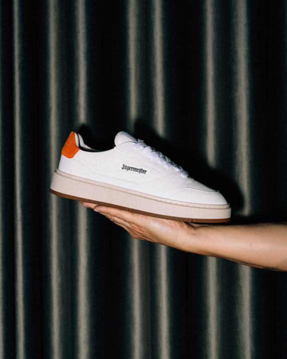A white Jägermeister sneaker with an orange heel tab and black Jägermeister wordmark, held in a person's hand against a dark vertically pleated curtain background.