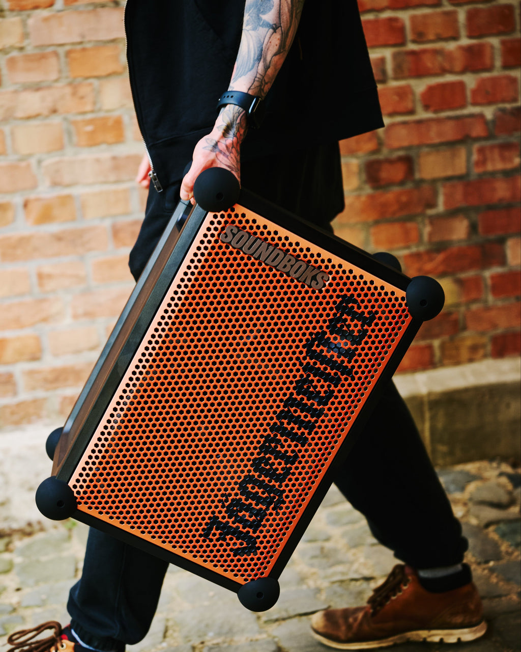 Egy személy narancssárga és fekete hordozható Soundboks hangszórót tart 'Jägermeister' márkajelzéssel egy téglafal előtt.