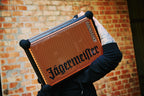 Egy személy narancssárga és fekete hordozható Soundboks hangszórót tart 'Jägermeister' márkajelzéssel egy téglafal előtt.