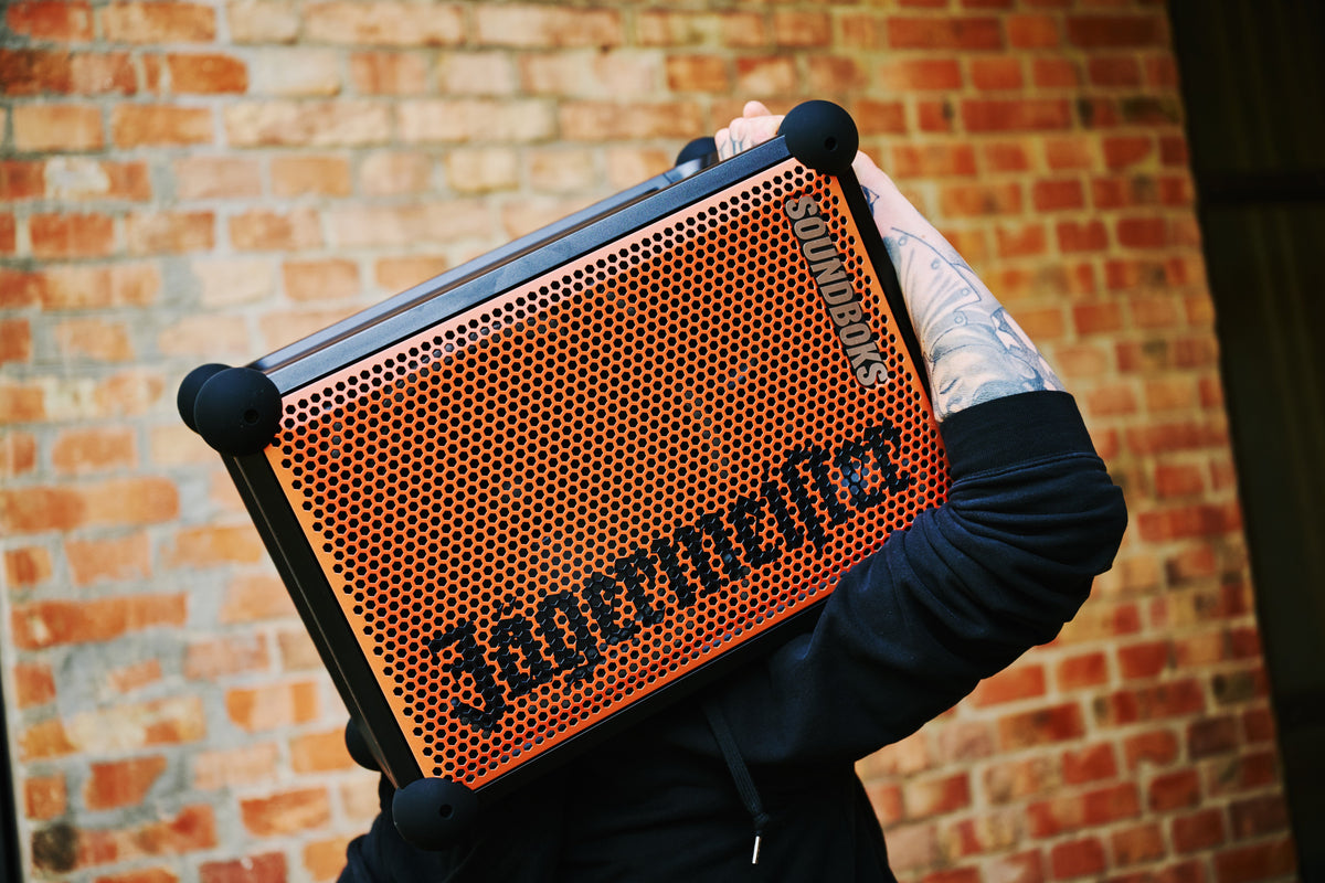 Egy személy narancssárga és fekete hordozható Soundboks hangszórót tart 'Jägermeister' márkajelzéssel egy téglafal előtt.