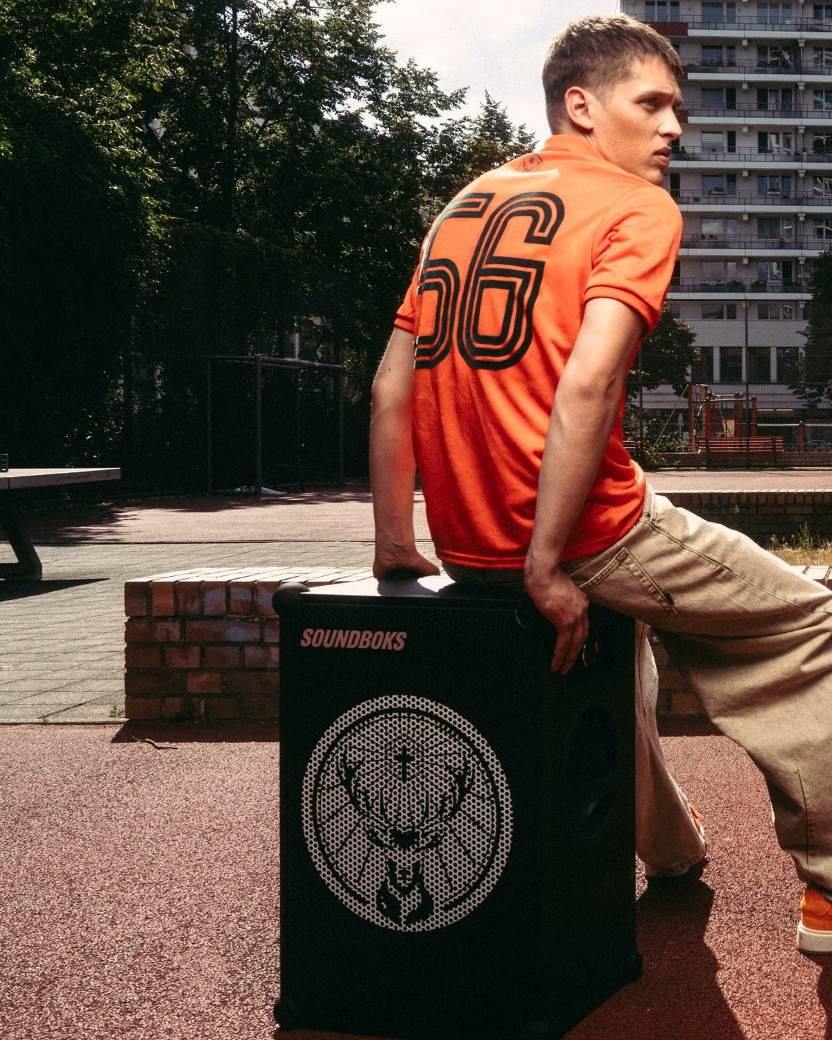 Egy férfi narancssárga Jägermeister mezt és bézs nadrágot visel, és egy fekete Soundboks hangszórón ül, Jägermeister márkalogóval