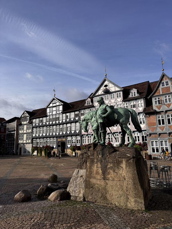A bronze equestrian statue stands on a large stone base in a town square in Wolfenbüttel, with historic half-timbered buildings and a blue sky in the background.