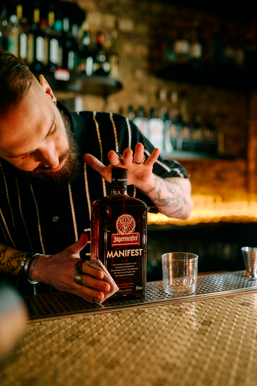 A bartender with a beard and tattoos leans over a bar counter, holding a bottle of Supermargaret Manifest whiskey. The bottle is positioned next to a clear rocks glass and a metal jigger, with a warm, blurred bar background.