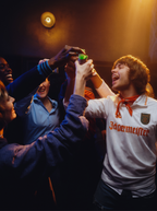 Emberek csoportja ünnepel egy Jägermeister pólót viselő személlyel egy félhomályos szobában.